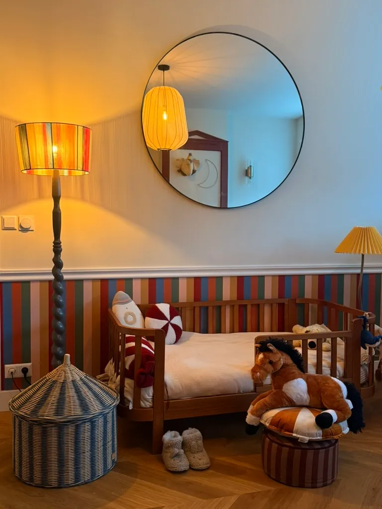 A child’s bed under a round mirror, lit by a striped floor lamp