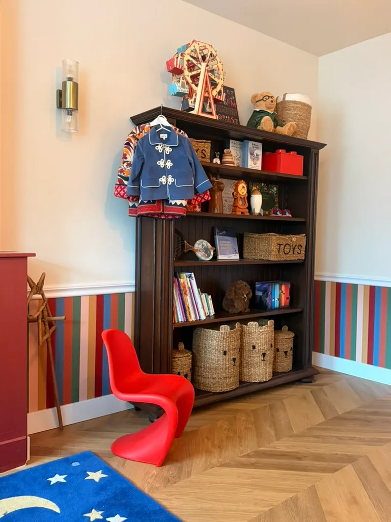 A dark wood bookshelf of books and woven toy baskets with a red moulded chair
